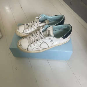  Philippe Model - Snygga vita sneakers från Philippe Model med svarta detaljer på hälen och ljusblå insida. Skorna har klassisk snörning, rund tå och platt sula. Tillverkade i skinn och passar perfekt till en avslappnad streetstyle-look. De är i bra skick förutom heelswipsen på ena skon det är storlek 41 men sitter som 42 hör av er vid minsta fundering