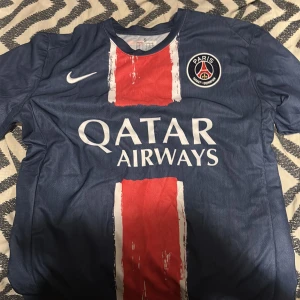 fotbollstshirt psg - Snygg blå Paris Saint-Germain fotbollströja från Nike med röd och vit randig design på framsidan. PSG-logga på bröstet och Qatar Airways tryck. Kortärmad och tillverkad i lätt, ventilerande material som passar perfekt för match eller träning.