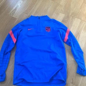 Blå långärmad Atlético Madrid Nike-tröja - Säljer en blå långärmad fotbollströja från Nike med Atlético Madrid-logga på bröstet och orange Nike-swoosh. Tröjan har dragkedja vid halsen och orange detaljer på ärmarna. Perfekt för träning eller match. Materialet är tunt och väldigt bekväm! Fullt äkta, och ruggigt snygg!