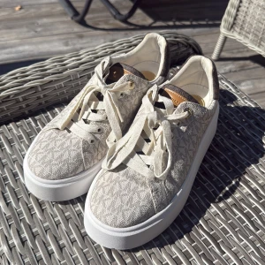 Beige sneakers från Michael Kors - Snygga sneakers från Michael Kors i beige med diskret logomönster och vita plattformssulor. Skorna har snörning med guldfärgade detaljer och bruna detaljer vid häl och plös. Tillverkade i textil och syntetmaterial, perfekta för en trendig vardagslook.