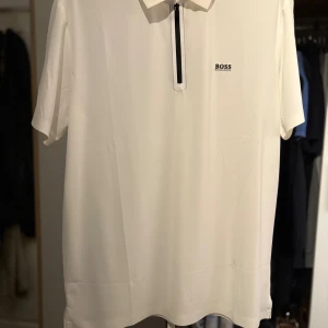 Vit kortärmad t-shirt från BOSS - Vit t-shirt från BOSS med korta ärmar och klassisk krage. T-shirten har en svart dragkedja framtill och diskret BOSS-logga på bröstet. Snygg och stilren design som passar perfekt till jeans eller shorts.