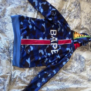 BAPE blå camo hoodie  - Säljer en blå camo hoodie från BAPE med shark-trycket på huvan och stora BAPE-loggan framtill. Hoodien har dragkedja, huva med färgglada detaljer och är tillverkad i mjukt material. 