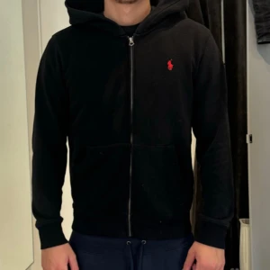 Svart hoodie från Polo Ralph Lauren - Svart hoodie från Polo Ralph Lauren med dragkedja och huva. Klassisk röd logga broderad på bröstet och känguruficka framtill. Tillverkad i mjuk bomull, perfekt för chill dagar. Snygg och enkel att matcha med allt.