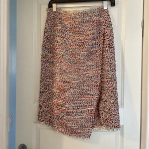 Flerfärgad omlottkjol från H&M - Säljer en snygg omlottkjol från H&M i storlek 38. Kjolen har ett vävt, flerfärgat tyg i vitt, blått och rött med fransig kant och asymmetrisk nederdel. Perfekt för dig som gillar unika detaljer och färg.