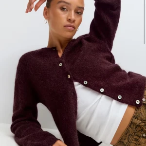 Vinröd alpaca kofta med knappar - Mysig mörklila kofta med lång ärm och snygga knappar framtill. Koftan är stickad och har en lite fluffig känsla, perfekt att slänga över en topp. Enkel och stilren design som passar till många outfits.