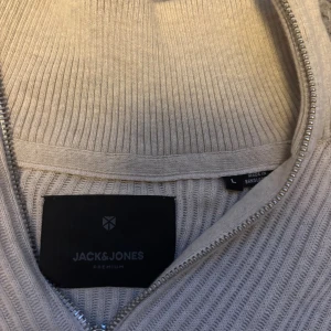 Beige ribbad half zip-tröja Jack & Jones - Ribbstickad beige tröja från Jack & Jones med half zip och hög krage. Långärmad modell i mjukt material, perfekt för dig som gillar stilren och enkel design. Snyggt strukturmönster och diskret logga på insidan av kragen. Pris kan diskuteras. Helt oanvänd.