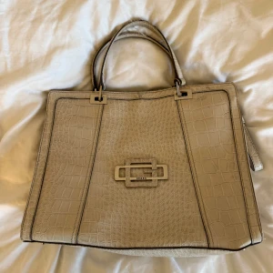 Beige handväska i croco-look från Guess - Snygg beige handväska från Guess med croco-mönstrad yta och stilren fyrkantig form. Väskan har dubbla handtag, dragkedja upptill och en metallogo framtill. Perfekt för dig som vill ha en trendig och rymlig väska med exklusiv känsla.