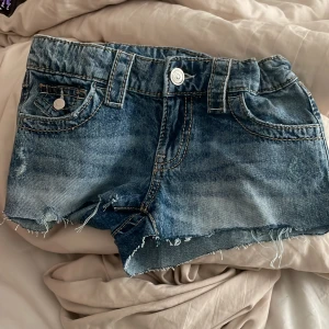 Blå jeansshorts från Gina Tricot - Snygga blå jeansshorts från Gina Tricot med råa kanter och slitna detaljer. Shortsen har klassiska fickor fram och bak med silverfärgade knappar och synliga sömmar. Perfekt för dig som gillar en chill och trendig stil.