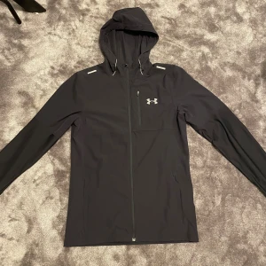 Svart vindjacka från Under Armour - Svart vindjacka från Under Armour med huva och snygga reflexdetaljer på axlarna. Jackan har dragkedja framtill, en bröstficka med zip och justerbar snörning vid huvan. Perfekt för dig som gillar sportig och clean stil.
