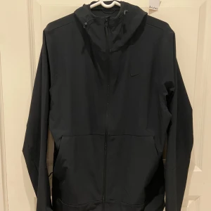 Svart Nike windbreaker  - Svart vindjacka från Nike med minimalistisk design, hel dragkedja framtill och justerbar huva med snörning. Jackan har diskret Nike-logga på bröstet och två sidofickor. Perfekt för blåsiga dagar och enkel att packa ner i väskan.