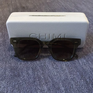 Gröna CHIMI solglasögon, fyrkantiga - Snygga solglasögon från CHIMI i en mörkgrön ton med fyrkantig form. Bågarna är tillverkade i blankt acetat och har två silverprickar på framsidan. Kommer med vit hård ask och mjuk påse med snörning. Perfekt för dig som vill ha en clean och modern look.