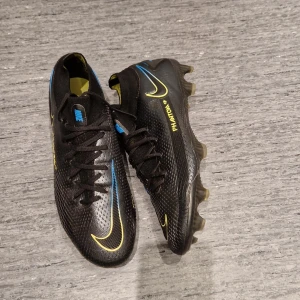 Nike Phantom GT2 elite Svarta Fotbollsskor - Säljer ett par Nike Phantom GT2 fotbollsskor i svart med gula och blå detaljer. Skorna har snörning, mönstrad ovandel i syntetmaterial och fasta dobbar för gräsplan. Snygg, sportig look med tydliga Nike-loggor och texten PHANTOM GT på sidan.