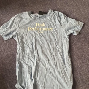 Ljusblå Peak Performance t-shirt - Ljusblå t-shirt från Peak Performance med klassisk rund hals och tryckt logga i vitt framtill. Skön passform och tillverkad i mjuk bomull, perfekt för en avslappnad stil. Enkel och stilren design som funkar till det mesta.