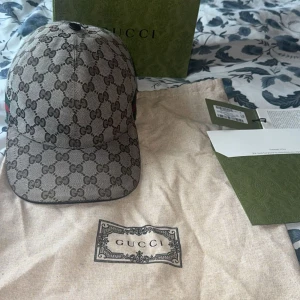 Gucci keps  - Snygg keps från Gucci med klassiskt GG-monogram i beige och brunt. Kepsen har en böjd skärm och justerbar rem bak. På sidan finns den ikoniska gröna och röda randen. Materialet är slitstark bomullscanvas med exklusiv känsla. Perfekt för dig som vill sticka ut med lyxig streetstyle. Tar emot byten 