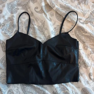 Svart faux skinn korsett-topp - Snygg svart korsett-topp i faux skinn med smala axelband och v-ringad skärning. 