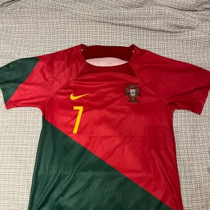 Portugal Ronaldo 7 fotbollströja Nike - Snygg Portugal fotbollströja från Nike med Ronaldo och nummer 7 på ryggen. Tröjan är röd med grönt diagonalblock, gula detaljer och landslagets emblem på bröstet. Kortärmad och tillverkad i lätt, ventilerande material – perfekt för match eller träning. Portugals 2022 World Cup tröja andvänd 1 gång på grund av att den var förstor för mig!