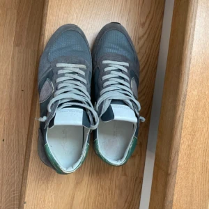 Philippe Model sneakers i grå mocka - Snygga Philippe Model sneakers i grå mocka och textil med gröna skinn-detaljer på hälen. Skorna har klassisk snörning och vit sula. Perfekta för dig som gillar en stilren men ändå sportig look.