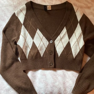 Brun croppad kofta från Divided XS - Superfin brun croppad kofta från Divided med klassiskt argylemönster i vitt och beige. Koftan har v-ringning, ribbade muddar och tre knappar framtill. Perfekt för lager-på-lager och ger en preppy vibe till din outfit.