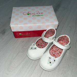 Vita ballerinaskor från Cupcake Couture, storlek 22 - Ett par söta vita ballerina skor från Cupcake Couture. Skorna har en blommig innersula och en justerbar rem över foten för en säker passform. Perfekt för speciella tillfällen eller för att piffa upp vardagsoutfiten.