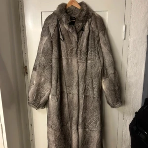 Lång grå pälsjacka i äkta päls - Säljer en lång och oversized pälsjacka i grå toner. Jackan har hög krage, breda ärmar och dragsko vid ärmsluten. Pälsen är mjuk och tät, perfekt för kalla vinterdagar. Fodrad insida för extra värme och komfort. Riktigt statement-plagg för dig som vill sticka ut.