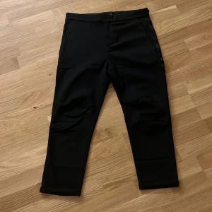 Svarta kostymbyxor Elias Rumelis - Svarta kostymbyxor från Elias Rumelis med croppad passform. Byxorna har raka ben, pressveck och snygga bakfickor med knappar. Perfekt för dig som gillar stilrena och moderna plagg. Materialet känns mjukt och har en lätt stretch.