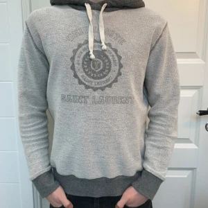 Grå hoodie från Saint Laurent - Säljer en snygg grå hoodie från Saint Laurent med tryckt logga och mönster framtill. Tröjan har kontrastfärgad mörkgrå huva, vita snören och ribbade muddar. Perfekt för dig som gillar exklusiva streetwear-plagg.
