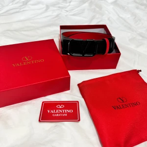 Valentino bälte - Snyggt svart skärp från Valentino Garavani med ikoniskt V-spänne i matt svart metall. Bältet har röda detaljer på insidan och vid hällan, vilket ger en cool kontrast. Tillverkat i äkta skinn och levereras med original ask, dustbag och kort.