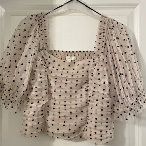 Prickig blus med puffärm från H&M - Supersöt ljusbeige blus från H&M med svarta prickar och transparenta puffärmar. Blusen har fyrkantig ringning, smockad rygg och voluminösa ärmar i mesh. Perfekt för dig som vill sticka ut med en trendig och lekfull look.
