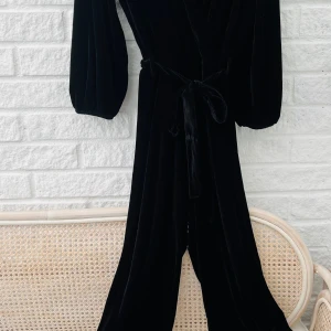 Svart sammetsbyxdress med knytband - Svart byxdress i mjuk sammet med vida ben och långa ärmar. Knytband i midjan ger en snygg siluett och plagget har en elegant känsla. Perfekt för dig som vill sticka ut med en cool och stilren look.