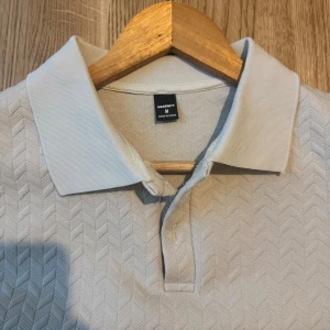 Beige pikétröja från Manfinity M - Snygg beige pikétröja från Manfinity i storlek M. Tröjan har klassisk krage, korta ärmar och ett diskret fiskbensmönster i tyget. Perfekt för en clean och stilren look. Materialet känns mjukt och bekvämt mot huden.