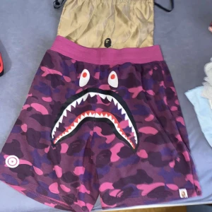 BAPE Shark Camo shorts lila/rosa - Säljer ett par BAPE Shark Camo shorts i lila och rosa med ikoniskt hajansikte framtill och tiger- och WGM-tryck baktill. Shorts med elastisk midja och camouflagemönster, tillverkade i mjuk bomull. Perfekt för dig som vill sticka ut med streetwear.