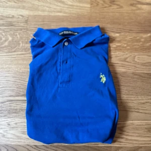Blå pikétröja från U.S. Polo Assn. - Klassisk blå pikétröja från U.S. Polo Assn. med korta ärmar och krage. Tröjan har knäppning framtill och den ikoniska broderade loggan i gult på bröstet. Tillverkad i mjuk bomull som känns skön mot huden. Perfekt för en clean och sportig stil.