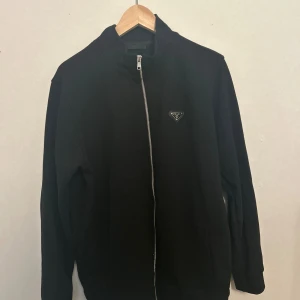 Svart bomberjacka från Prada - Svart bomberjacka från Prada med stilren design och rak passform. Jackan har en silverfärgad dragkedja framtill, ribbade muddar och ett diskret Prada-emblem på bröstet. Tillverkad i 100% bomull för en skön och lyxig känsla. Perfekt för dig som gillar exklusiva streetwear-vibbar.