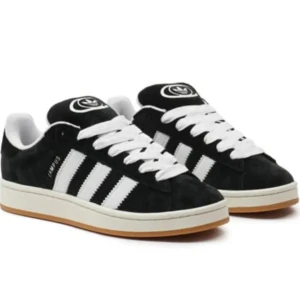 Adidas Campus svarta sneakers mocka - Svarta Adidas Campus sneakers i mocka med klassiska vita ränder på sidorna och vit sula. Skorna har rund tå, platt sula och vita skosnören. Perfekta för en stilren och sportig look. 