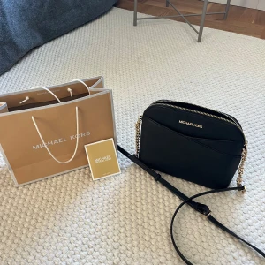 Svart axelväska från Michael Kors - Snygg svart axelväska från Michael Kors i skinn med guldfärgade kedjedetaljer och justerbar axelrem. Väskan har dragkedja upptill och ett fack på insidan. Helt oanvänd, helt ny i perfekt skick. Nypris: 3300. Fått som present, köpt i butik.