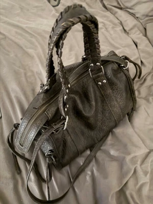 Svart axelväska med flätade handtag - Säljer en svart axelväska i skinn med snygga flätade handtag och silverfärgade metalldetaljer. Väskan har dragkedjor både upptill och på sidorna samt långa remmar med knyt. Perfekt storlek för allt du behöver ha med dig och riktigt cool design.