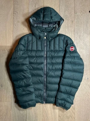 Mörkgrön pufferjacka från Colmar - Säljer en mörkgrön pufferjacka från Colmar med huva och snygga quiltade paneler. Jackan har dragkedja framtill och två tryckknappar vid halsen. Perfekt för kalla dagar och har en clean look med Colmar-logga på ärmen. Storlek 44 S