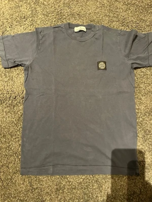 Mörkblå Stone Island Junior t-shirt - Säljer en mörkblå t-shirt från Stone Island Junior med klassisk rund hals och korta ärmar. T-shirten har det ikoniska Stone Island-märket broderat på bröstet och är tillverkad i mjuk bomull. Perfekt för dig som gillar streetwear och snygga loggor.