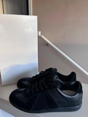 Svarta Maison Margiela sneakers - Maison Margiela sneakers helt nya och använda  i helsvart med en mix av mocka och skinn. Klassisk låg modell med rund tå, snörning och platt sula. Snygg minimalistisk design som passar till allt. Kommer med originalkartong och dustbags.