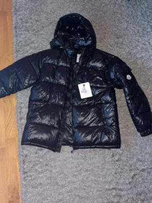 Svart Moncler pufferjacka med luva - Svart pufferjacka från Moncler med glansig finish och huva. Jackan har dragkedja framtill, två sidofickor och Moncler-logga på ärmen. Insidan har ett coolt serietryck. Perfekt för kalla dagar och riktigt snygg streetstil. 