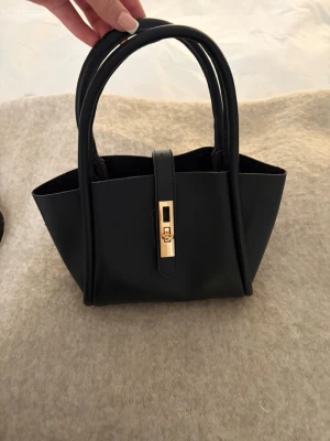 Svart handväska med gulddetalj - Stilren svart handväska i skinn med två handtag och en snygg guldfärgad låsdetalj framtill. Väskan har en rymlig insida och medföljande axelrem som kan fästas. Väskan är aldrig använd så väldigt fint skick, skriv privat för fler bilder eller om du undrar något!🩷