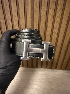 Svart skärp med stor H-spänne - Säljer ett svart skärp med en rejäl metallspänne i form av ett H. Skärpet har vita kontrastsömmar längs kanterna och är gjort i slätt skinn. Perfekt accessoar för att lyfta vilken outfit som helst.