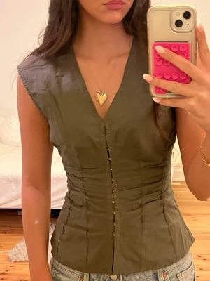 Grön korsettopp från Weekday - Säljer en khaki/mörkgrön korsettopp från Weekday i storlek 36. Toppen har en v-ringad design, är ärmlös och har snygga knappar framtill. Materialet är mjukt och lätt, perfekt för att styla med jeans eller kjol. Figurnära passform som framhäver midjan.