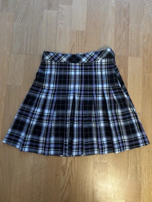 Rutig kortkjol från Lindex - Säljer en snygg rutig kortkjol från Lindex i svart, vit och lila. Kjolen har klassiskt veckad design, knapp och dragkedja i sidan för enkel påklädning och har justerbart resårband i midjan.