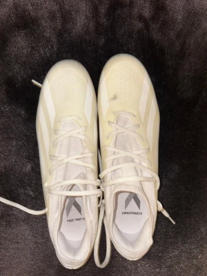 Vita adidas Crazyfast.3 fotbollsskor - Säljer ett par vita adidas Crazyfast.3 fotbollsskor med klassiska tre ränder på sidorna. Skorna har snörning och en ovandel i syntetmaterial för lätt känsla och bra bollkontroll. Yttersulan har fasta dobbar för spel på gräs. Perfekta för dig som vill ha speed på planen.