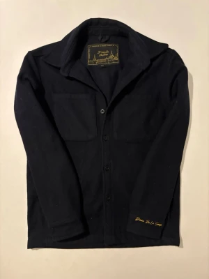 Svart overshirt från D'once De La Sense - Svart overshirt i 100% ull från D'once De La Sense. Jackan har klassisk krage, två stora bröstfickor och knäppning med svarta knappar framtill. Snygg broderad detalj på ärmen och stilren design som funkar till många outfits.