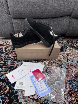 Svarta sneakers från Christian Louboutin - Säljer ett par svarta sneakers från Christian Louboutin i mocka med klassisk röd sula och diskret logga på sidan. Skorna har rund tå, snörning och platt sula. Medföljer originalkartong och äkthetsbevis. Perfekt för dig som gillar lyxiga detaljer och stilren design. Skobox ingår! Priset kan diskuteras
