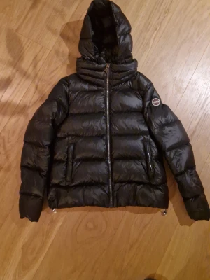 Svart pufferjacka från Colmar - Säljer en svart pufferjacka från Colmar med huva och snygga horisontella sömmar. Jackan har dragkedja framtill och två fickor med dragkedja. Perfekt för kalla dagar och har en cool, glansig finish som ger extra edge till din stil.