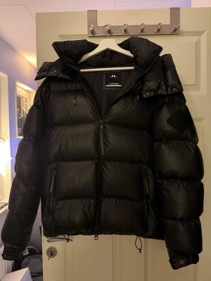 Svart pufferjacka från J.Lindeberg - Säljer en svart pufferjacka från J.Lindeberg med hög krage och huva. Jackan har stora, quiltade sektioner, dragkedja framtill med J.Lindeberg-tryck. Jackan är i stort sett som ny! Perfekt för kalla dagar och har en riktigt snygg, modern look. Passar om du är ca 175-180 cm lång vid snabb affär är priset diskuterbart