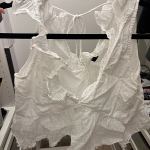 Vit volang linnetopp med knyt - Säljer en söt vit linnetopp med volangdetaljer och knyt i ryggen. Toppen är ärmlös och har en luftig, lätt struktur som passar perfekt till varma dagar. Den har en omlottliknande design framtill och romantiska volanger längs kanterna.